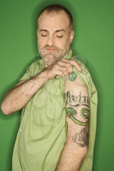 Portrait of Caucasian man pulling up sleeve to show tattoo standing against green background.