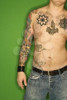 Bare torso of Caucasian man with tattoos standing against green background.