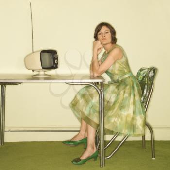 Pretty Caucasian mid-adult woman wearing green vintage dress sitting at 50's retro dinette in front of old televsion looking bored.