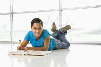Asian preteen girl lying on floor doing homework looking up at viewer.