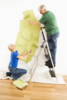 Middle-aged couple painting wall green with male on ladder.