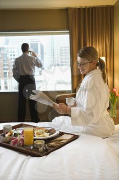 Caucasian woman in a robe sits on a hotel bed while reading the newspaper. A man stands in the background talking on his mobile phone. Vertical shot.