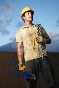 Male Caucasian construction worker wearing a yellow hardhat stands with heavy chain on his shoulder and bolt cutters in his hand. Vertical shot.