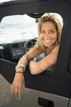 Portrait of a smiling young Caucasian female sitting in the driver's seat of an SUV leaning out the window with ocean waves in the background. Vertical format.