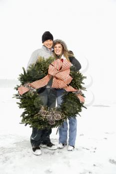 Young adult couple in the winter holding a Christmas wreath and with arms around one another. Vertical shot.