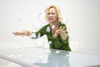 Caucasian middle aged businesswoman sitting at office desk throwing ripped up paperwork looking disgruntled.