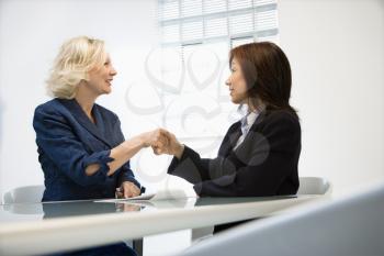 Two businesswomen sitting at office desk shaking hands.