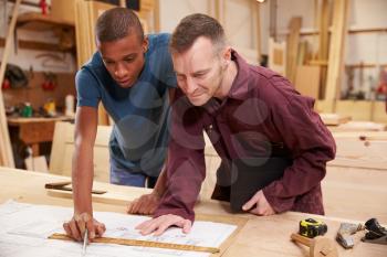 Carpenter With Apprentice Looking At Plans In Workshop