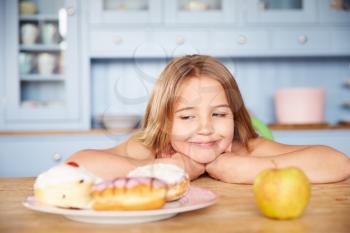 Girl Sitting At Table Choosing Cakes Or Apple For Snack