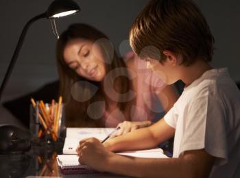 Teenage Sister Helping Younger Brother With Studies At Desk In Bedroom In Evening