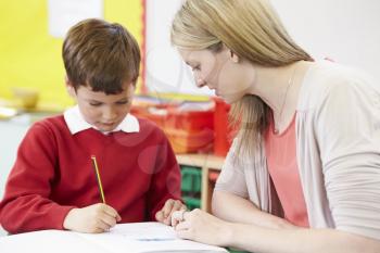 Teacher Helping Male Pupil With Practising Writing At Desk