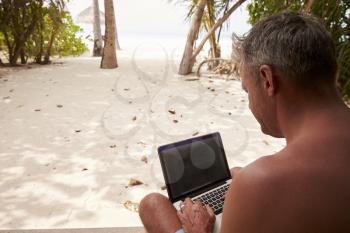 Man using a laptop computer on a beach, over shoulder view