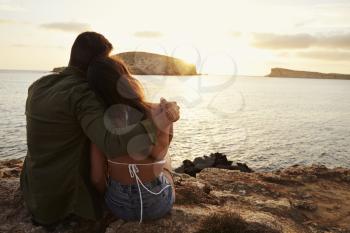 Rear View Of Couple Sitting On Cliff Watching Sunset