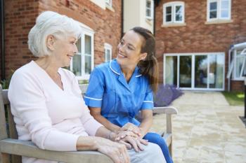 Senior Woman Sitting On Bench And Talking With Nurse In Retirement Home