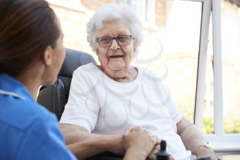 Senior Woman Sitting In Motorized Wheelchair Talking With Nurse In Retirement Home