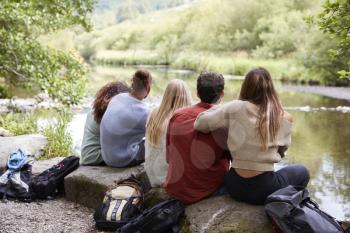 Five young adult friends taking a break sitting on rocks by a stream during a hike, back view, close up