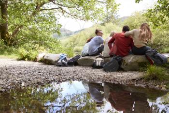 Five young adult friends taking a break sitting on rocks by a stream during a hike, back view
