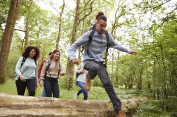 Multi ethnic group of five young adult friends running in a forest and jumping over fallen tree during a hike