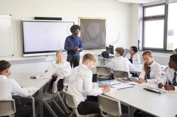 Male High School Teacher Standing Next To Interactive Whiteboard And Teaching Lesson To Pupils Wearing Uniform