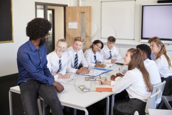 Male High School Teacher Sitting At Table With Teenage Pupils Wearing Uniform Teaching Lesson