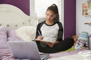 Teen girl sitting on bed watching computer and writing notes