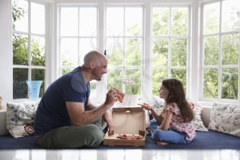 Dad and daughter sit on window seat at home sharing a pepperoni pizza