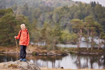 A boy standing on a rock by a lake holding a stick, smiling to camera, Lake District, UK