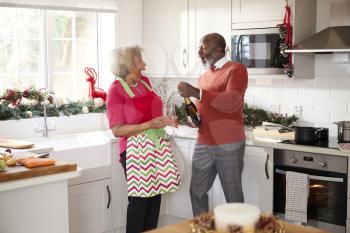 Senior black couple opening champagne and laughing together in the kitchen while preparing dinner on Christmas Day, selective focus