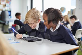 Two primary school kids sitting together at desk in a classroom using a tablet computer and stylus, close up
