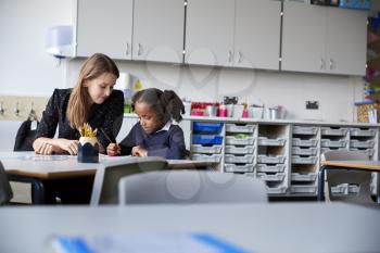Young female primary school teacher working one on one with a schoolgirl sitting at table in a classroom, selective focus