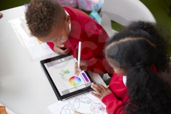 Elevated view of two kindergarten school kids sitting at a desk in a classroom drawing with a tablet computer and stylus, close up