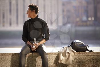 Young black woman wearing turtleneck sweater sitting in the sun on an embankment wall by the River Thames in London, backlit