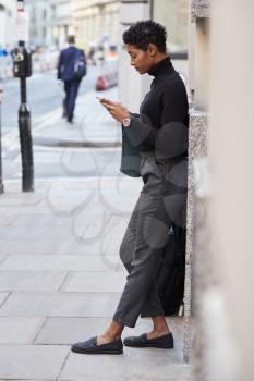 Young adult woman leaning on a wall in a London street using her smartphone, full length