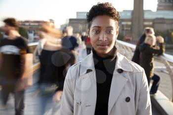 Young black woman standing on Millennium Bridge, London, looking to camera while pedestrians pass by, motion blur