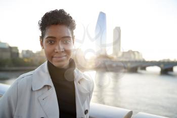 Young black woman wearing a turtleneck sweater and a mackintosh standing on Millennium Bridge, London, looking to camera smiling