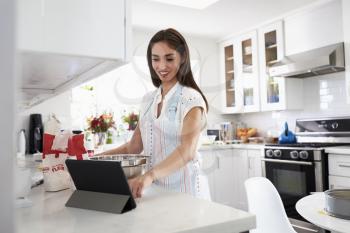Millennial Hispanic woman preparing cake mixture in the kitchen, using a recipe on a tablet computer
