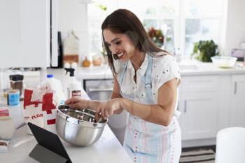 Smiling millennial woman preparing cake mixture, following a recipe on a tablet computer