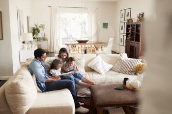 Young Hispanic family sitting on sofa reading a book together in the living room, seen from doorway