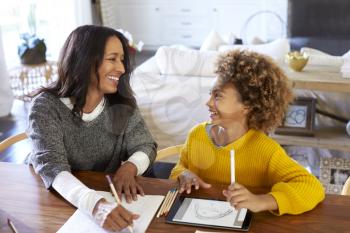 Middle aged mixed race woman sitting at a table in the dining room looking at her granddaughter, who is drawing using a tablet computer, elevated view, close up