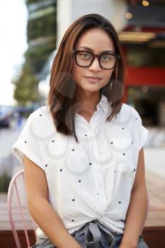 Trendy young Asian woman wearing glasses sitting on chair in the street looking to camera, close up