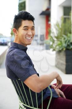 Millennial Asian man sitting on a chair in a city street, turning and smiling to camera, close up