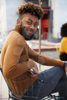 Millennial black hipster man sitting outside a cafe in the street, turns smiling to camera, close up