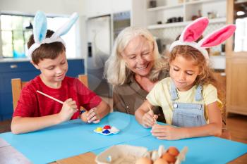Grandmother With Grandchildren Wearing Rabbit Ears Decorating Easter Eggs At Home Together