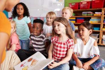 Group Of Elementary School Pupils Sitting On Floor Listening To Female Teacher Read Story