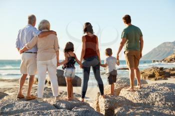 Three generation white family on a beach stand holding hands, admiring view, full length, back view