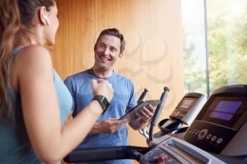 Woman In Gym With Personal Trainer Analysing Performance Using Smart Watch And Digital Tablet