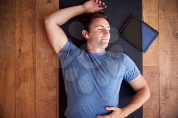 Overhead View Of Man Lying On Exercise Mat With Digital Tablet Listening To Wireless Headphones