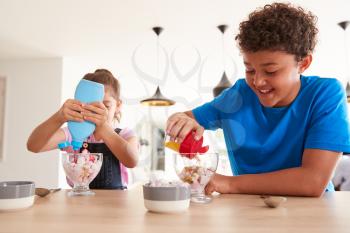 Children In Kitchen At Home Adding Sprinkles And Sauce To Ice Cream Dessert