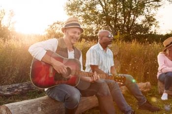 Group Of Mature Friends Sitting Around Fire And Singing Songs At Outdoor Campsite