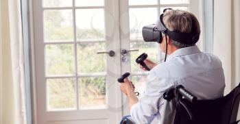 Mature Disabled Man In Wheelchair At Home Wearing Virtual Reality Headset Holding Gaming Controllers